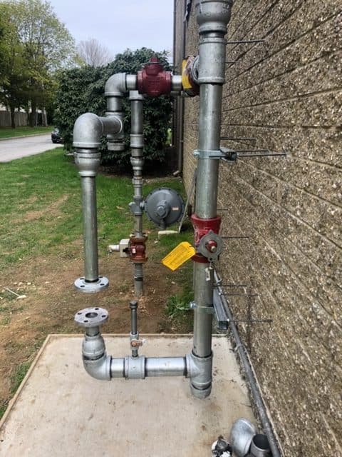 The pipes and valves attached to the building's exterior are part of an outdoor utility setup, possibly maintained by a skilled plumber. Red and yellow valve handles and secured metal brackets detail the system, with grass and a road in the background.