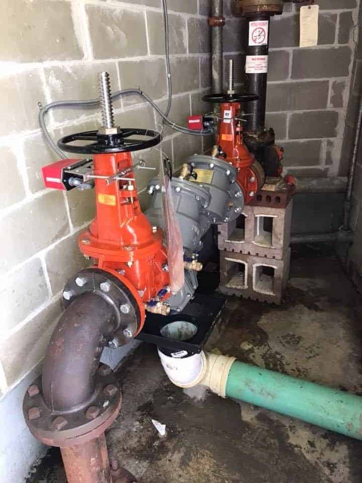 A close-up of industrial water pipes with red valves in a building showcases a setup perfect for Oil to Gas Conversions. The large metal pipe connects to a green one, supported by cinder blocks against concrete block walls. A warning sign is visible in the background.