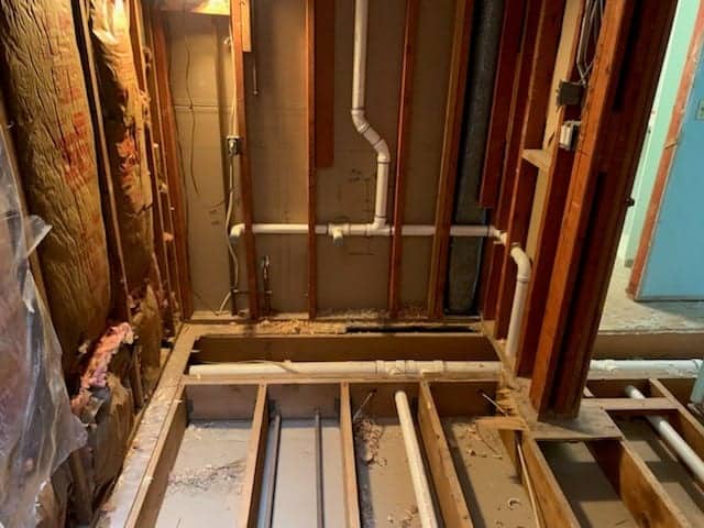 A room under renovation with exposed wooden beams and electrical wires awaits a residential plumber. Walls stripped to reveal insulation, pipes, and potential for mini split AC installations. The floor partly removed shows joists and subfloor, while plastic sheeting covers sections under dim ceiling light.