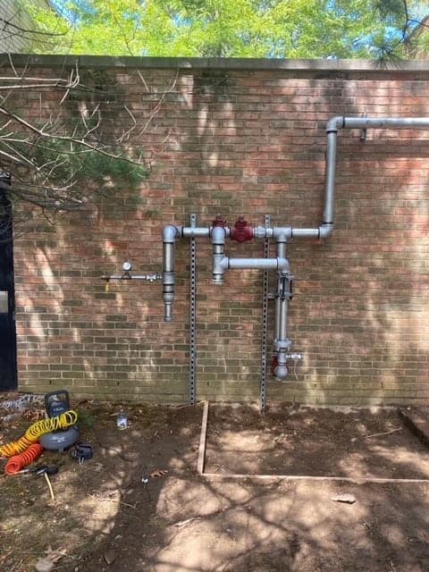 Pipes and valves are mounted on a brick wall outside, hinting at recent boiler services. A coil of orange and yellow hose lies alongside tools on the ground below, possibly left by a residential plumber. The scene is partially shaded by nearby trees.