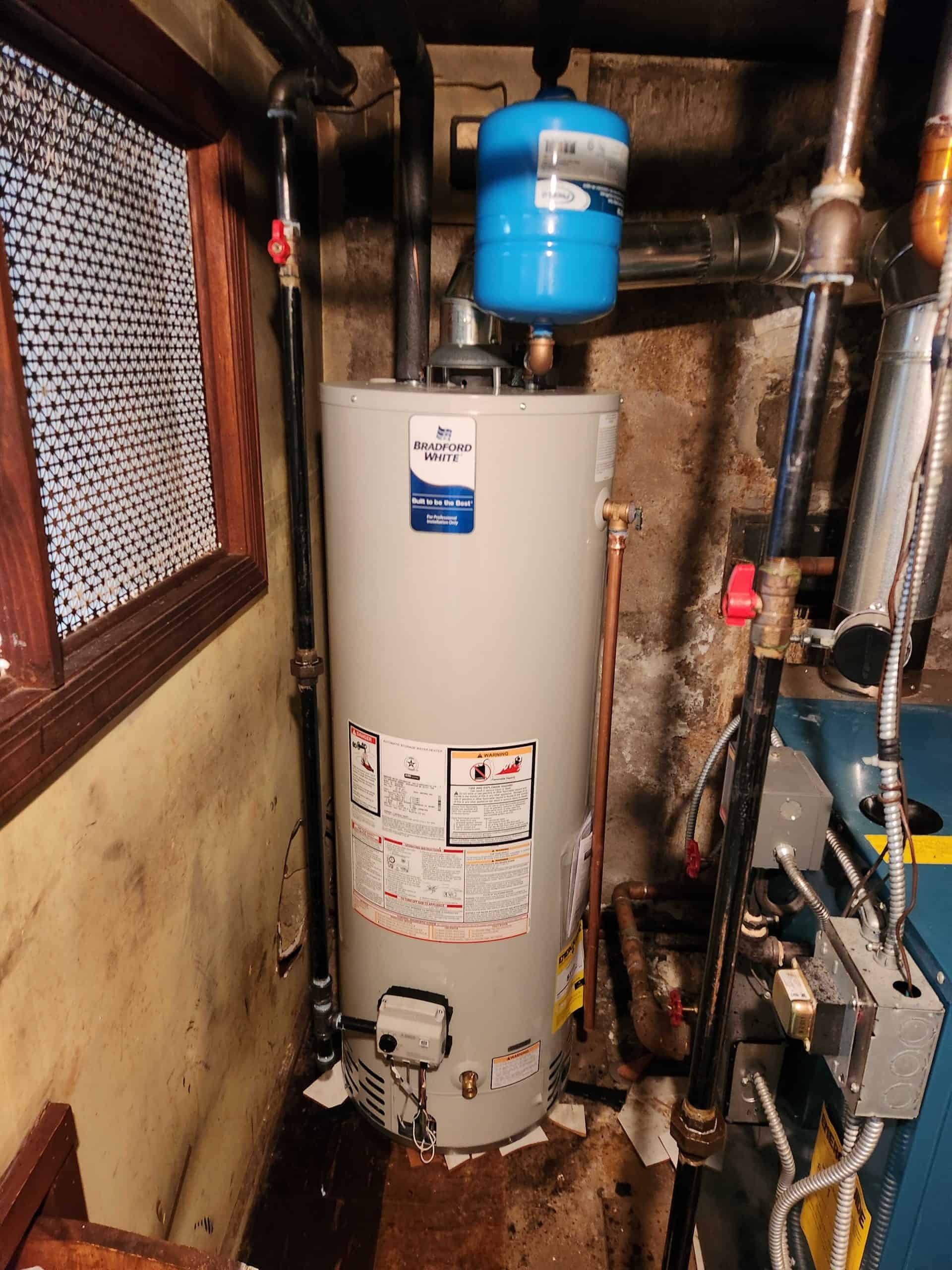 A water heater is installed in a basement corner, with various pipes connected. An expansion tank is mounted above. The yellowed walls and wooden floor suggest age. Nearby, a window with wire mesh lets in light. Expert boiler services ensure everything runs smoothly and efficiently.