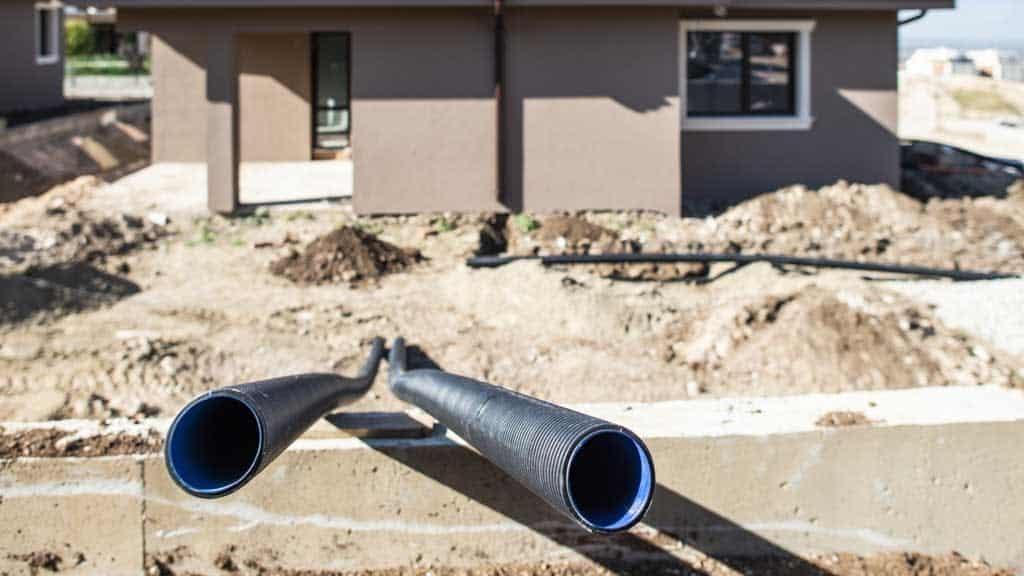 Two black pipes protrude from a concrete slab at a construction site, where dirt and building materials are scattered around. A residential plumber works nearby, ensuring everything is ready for mini split AC installations. In the background, a partially built brown building rises under a clear sky.