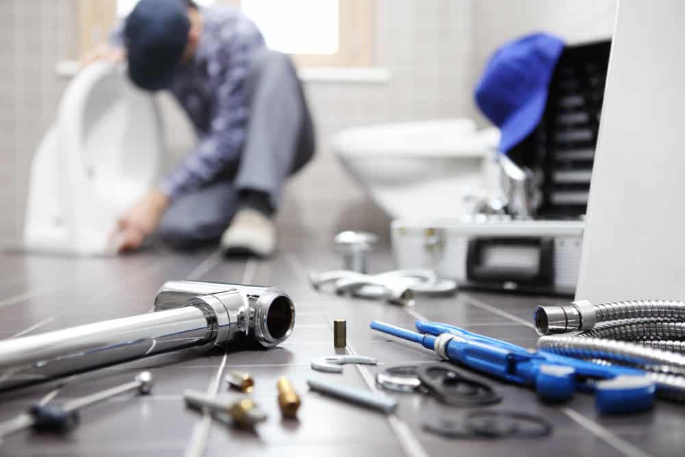 A residential plumber is fixing a toilet in a bathroom, tools scattered on the tile floor. The focus is on wrenches, bolts, and pipes, with a toolbox and a blue cap nearby—a testament to his expertise in plumbing and boiler services.
