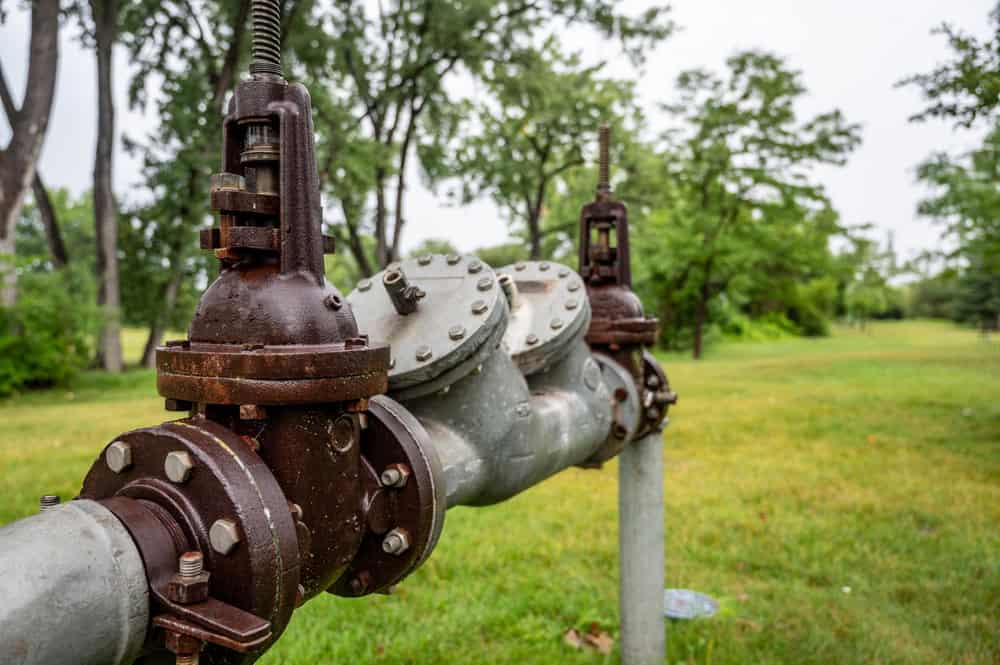 A close-up of a large rusted pipe with valves in a grassy park setting hints at the attention needed by a skilled plumber. Tall trees loom in the background on an overcast day, evoking the peaceful resilience found in expert residential plumbing services.