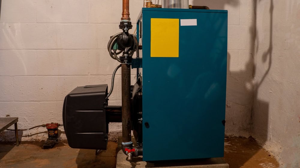 A blue furnace unit with pipes and a yellow sticker is installed against a beige basement wall on a concrete floor. Shadows are cast on the wall, and part of a rusted can and pipes can be seen in the background, hinting at recent upgrades by a residential plumber familiar with oil to gas conversions.