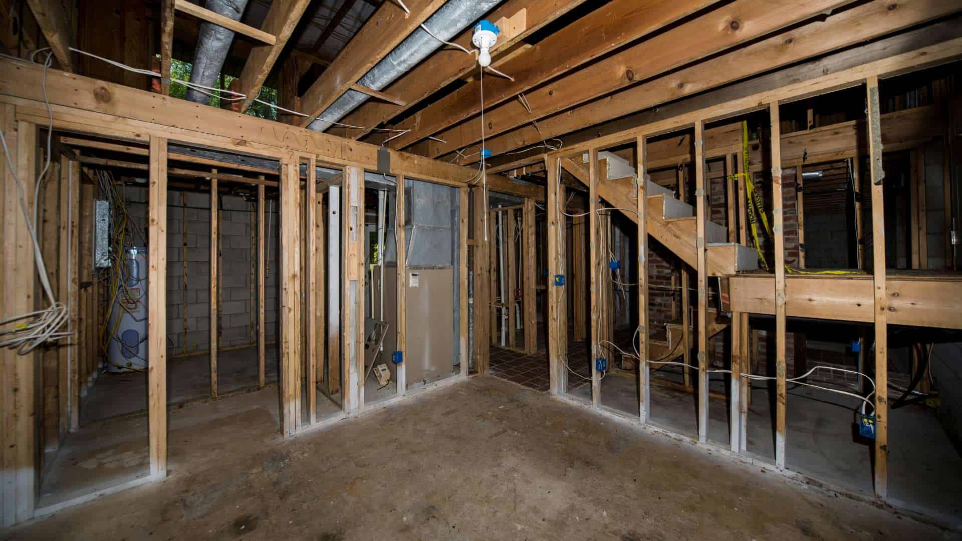 An unfinished basement with exposed wooden framing, concrete floor, and visible ductwork and wiring, ready for enhancements like a mini split AC installation. Stairs lead to an upper level; no drywall or flooring is installed yet.
