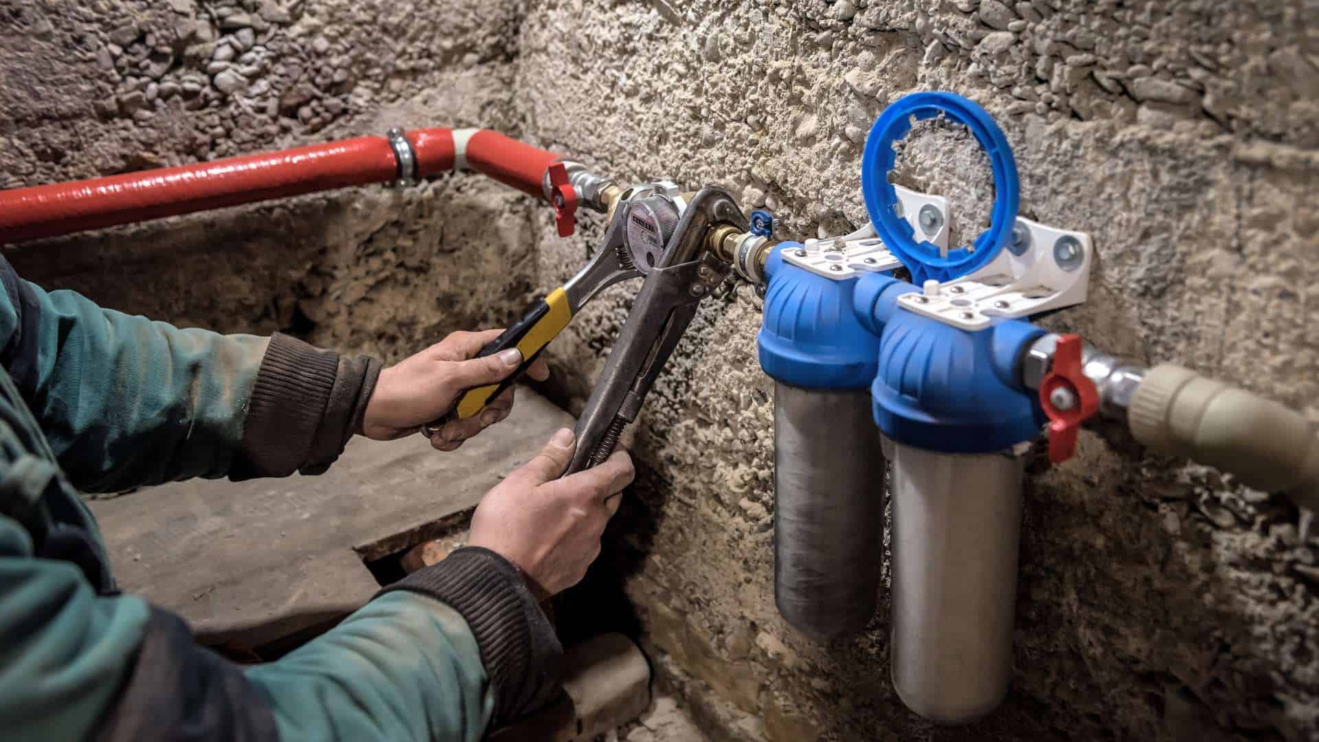 A plumber uses a wrench to install or adjust water filtration cartridges attached to a stone wall. The system, with its dual blue and white filters, features red pipes expertly connected by the plumber.