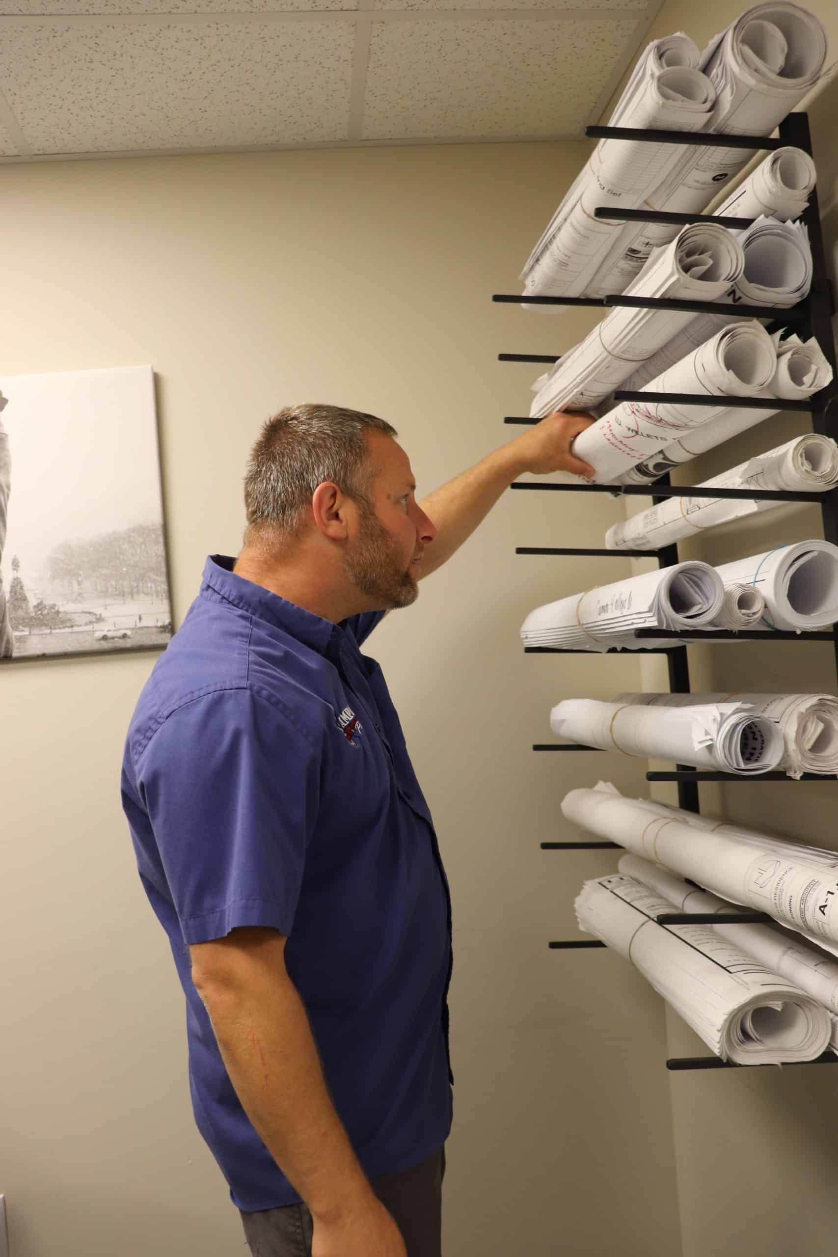 A man in a blue shirt reaches for a rolled-up architectural blueprint from a wall rack filled with similar blueprints, planning an oil to gas conversion project in an office setting.
