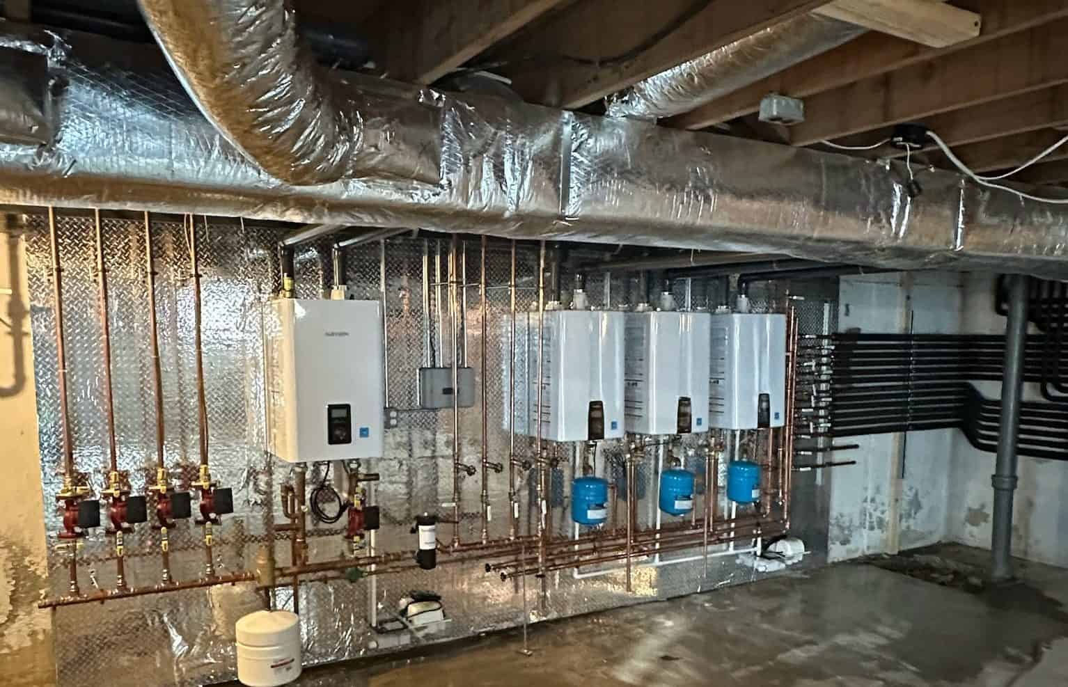 A basement with three water heaters mounted on the wall, surrounded by an intricate network of copper pipes. The ceiling is unfinished, featuring exposed wooden beams and insulated ducts. Perfect for a residential plumber handling Boiler Services, the floor remains bare concrete.