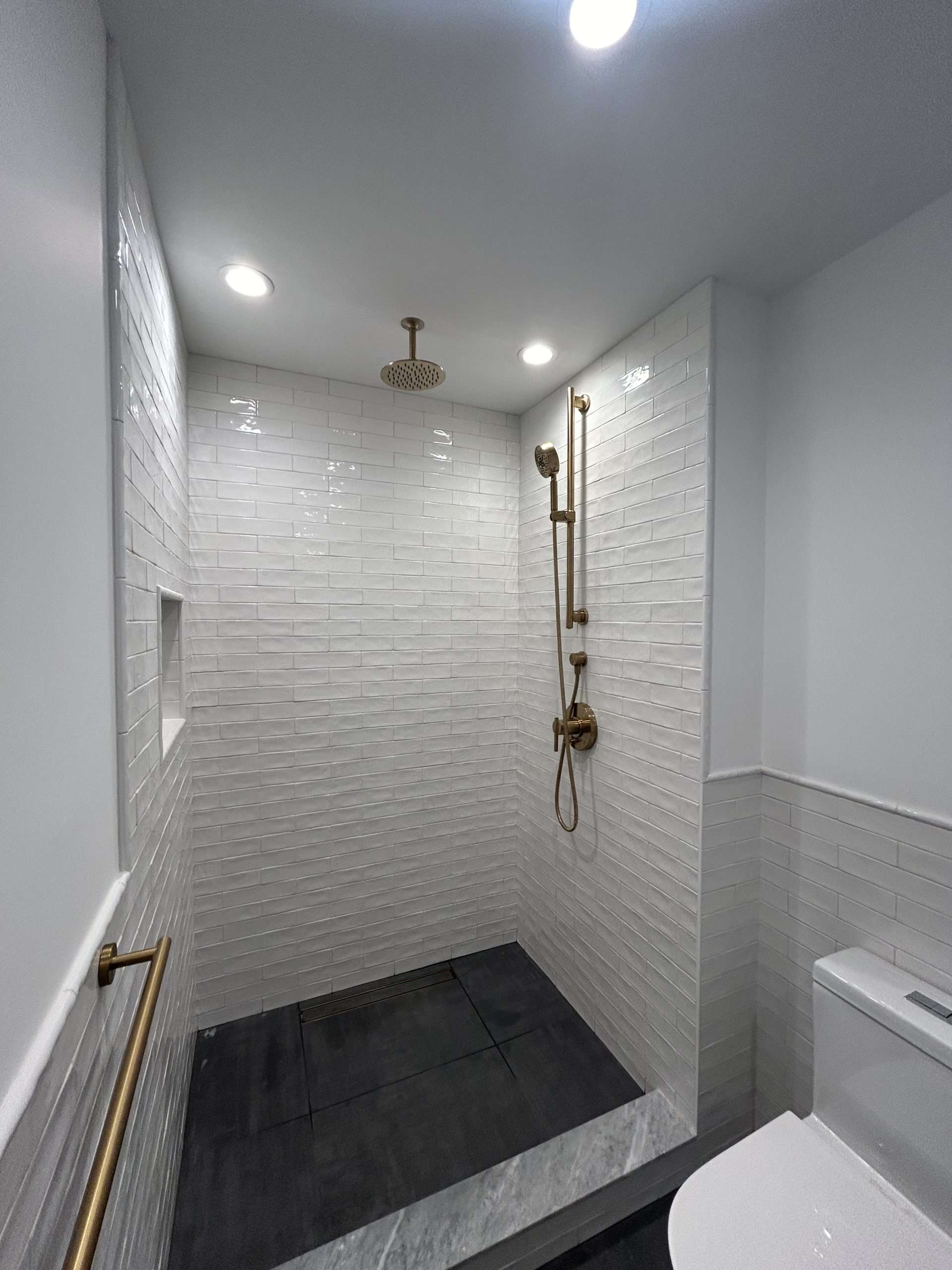 A modern bathroom designed by a skilled residential plumber features a spacious shower with white subway tiles and a brass rainfall showerhead. The dark gray tile floor complements the built-in niche, while recessed lighting brightens the space, complete with a stylish brass handrail.