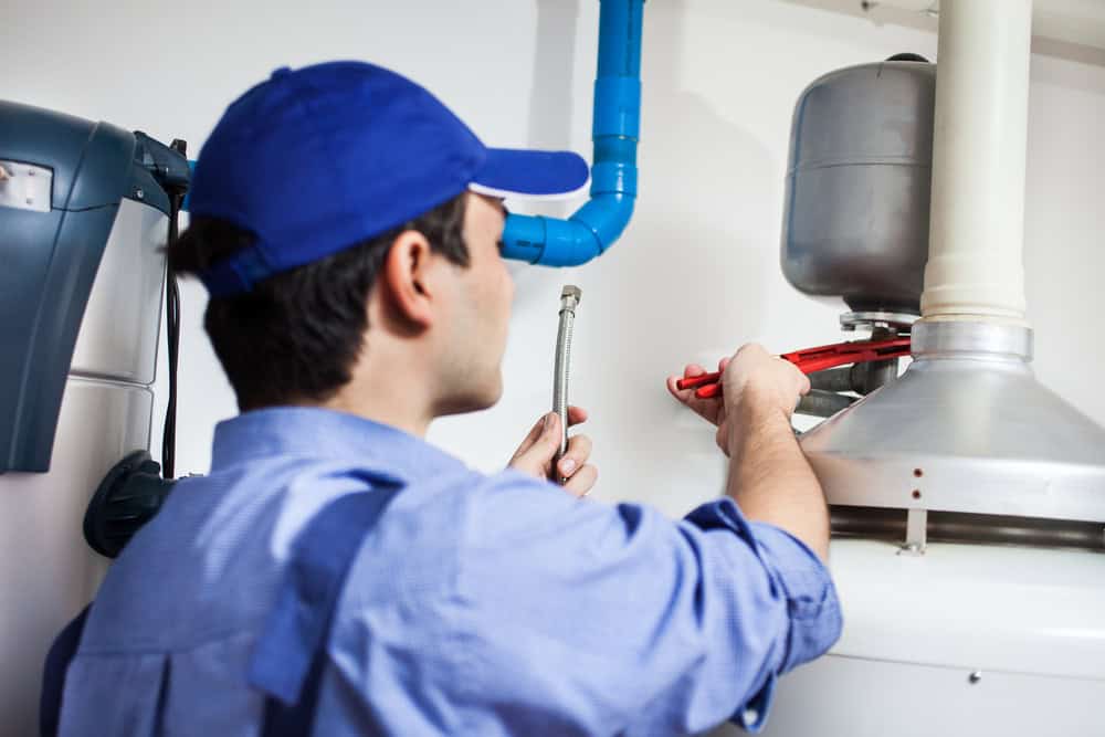 A plumber in a blue uniform and cap is using a red wrench to adjust a metallic pipe connected to a water heater. Blue and white pipes are visible in the background, highlighting expert boiler services at work.