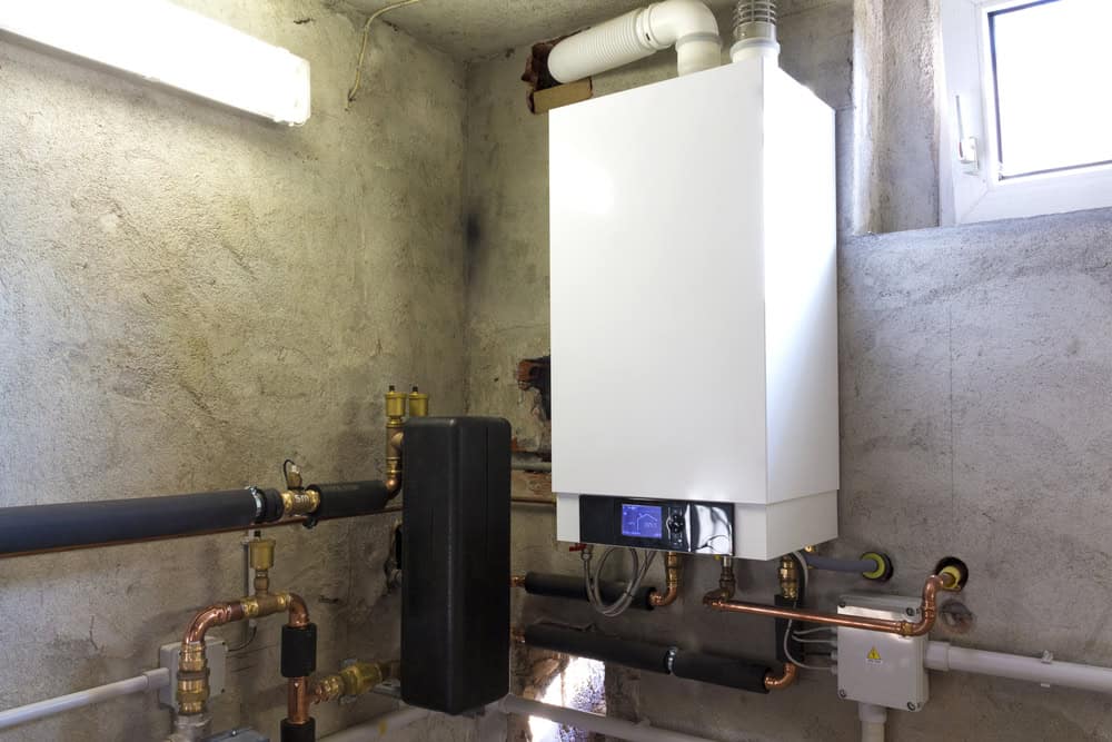 A modern boiler system mounted on a textured concrete wall in a utility room, surrounded by various pipes and fittings. A small window above lets in natural light, highlighting the precision of Residential Plumber craftsmanship.