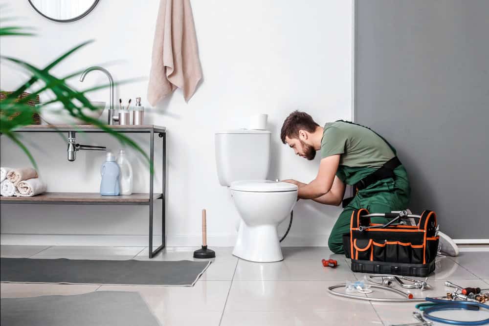 A skilled plumber in green overalls is kneeling while fixing a toilet in a clean, modern bathroom. Nearby, a tool bag and various tools are scattered on the floor. In the background, a sink with towel and toiletries adds to the tidy atmosphere as he discusses mini split AC installations with the homeowner.