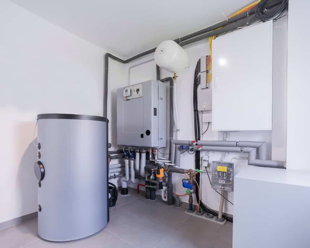 A modern boiler system installed in a basement by expert Boiler Services, featuring pipes, valves, a cylindrical water tank, and control panels. The setup is neatly arranged against a white wall on a tiled floor.