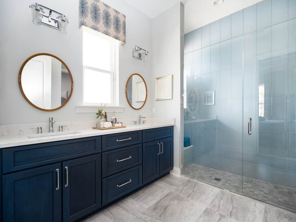 Modern bathroom with a double sink vanity featuring dark blue cabinets and round mirrors. A large glass-enclosed shower is on the right. The floor is light gray tile, and a window above the sinks allows natural light into the space, while a sleek mini split AC installation ensures comfort year-round.