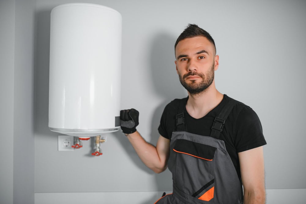 A plumber in work overalls stands next to a white water heater mounted on a gray wall. He has short dark hair and a beard, wearing a black shirt and gloves. His left hand rests on the heater, showcasing his expertise in boiler services.