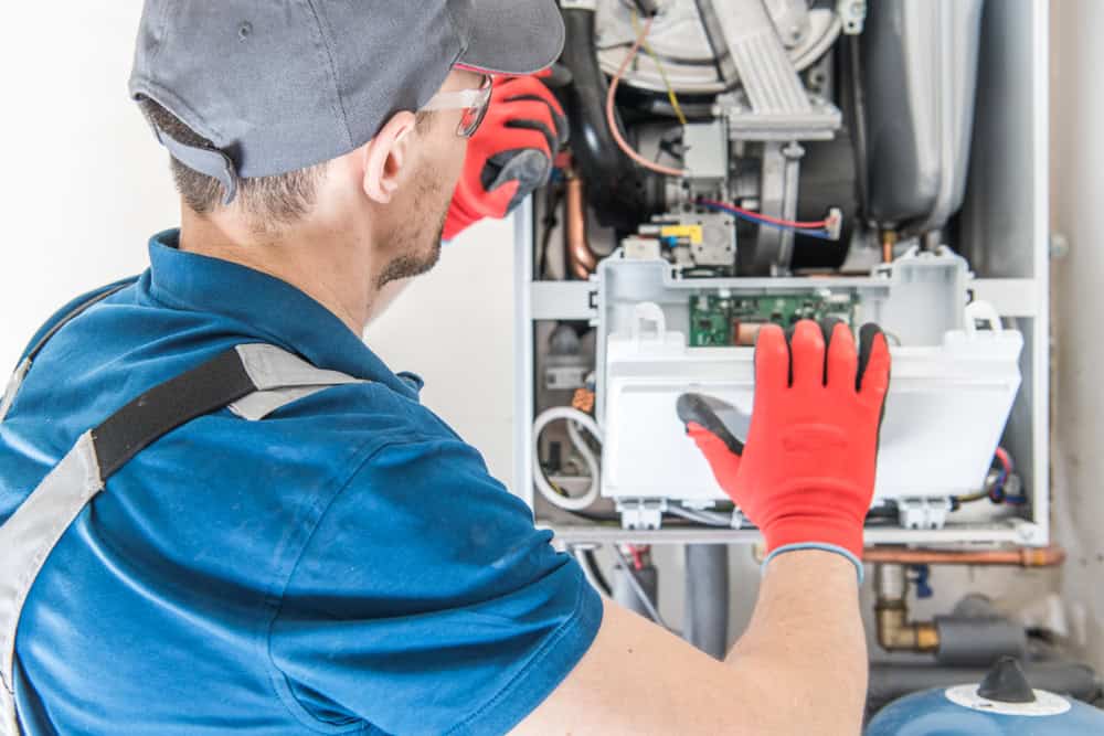 A maintenance technician in a blue uniform, red gloves, and cap meticulously inspects or repairs a gas furnace's internal components. Specializing in oil to gas conversions, he uses tools to deftly work on the exposed circuit board within the unit.