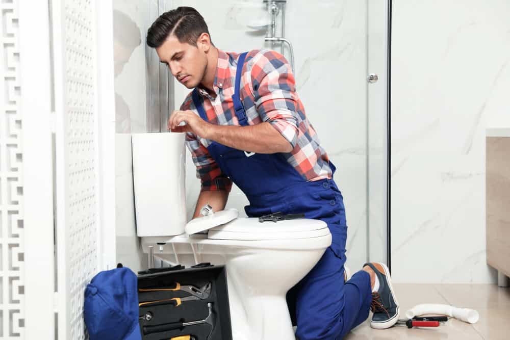 A residential plumber in blue overalls and a plaid shirt kneels beside a toilet, working on plumbing repairs. A tool bag and various tools are on the floor next to him in a bright, tiled bathroom.