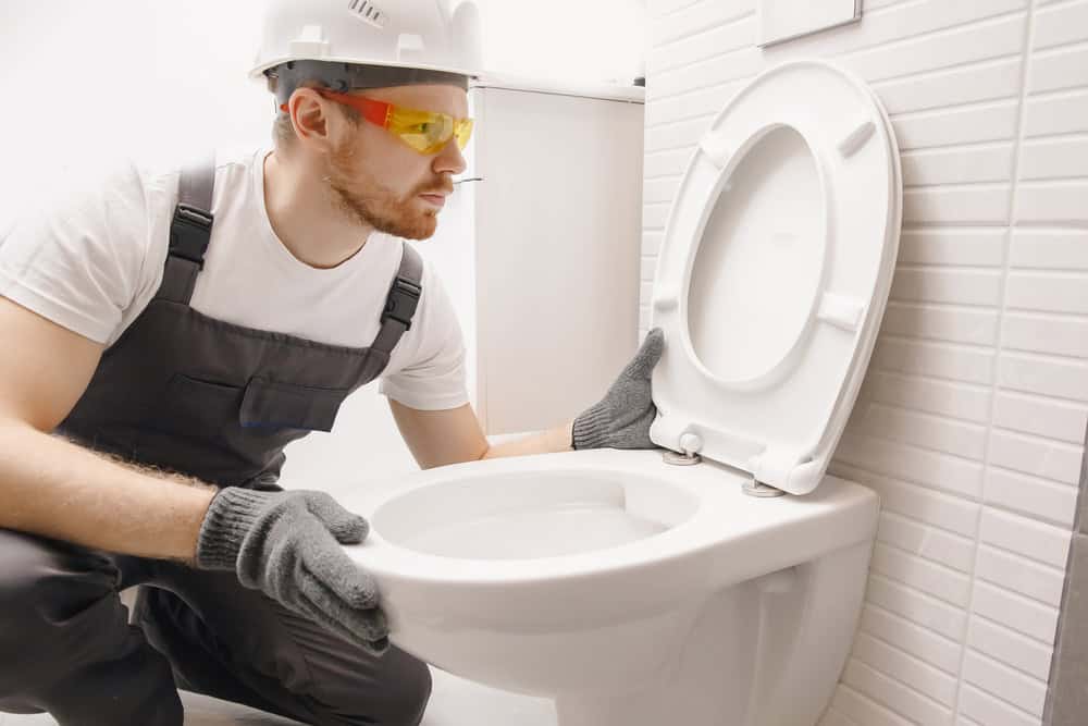 A plumber wearing a hard hat, safety glasses, and gloves inspects a white toilet seat in the bathroom. Kneeling in dark coveralls, he’s ready for any task, from simple repairs to complex projects like mini split AC installations.