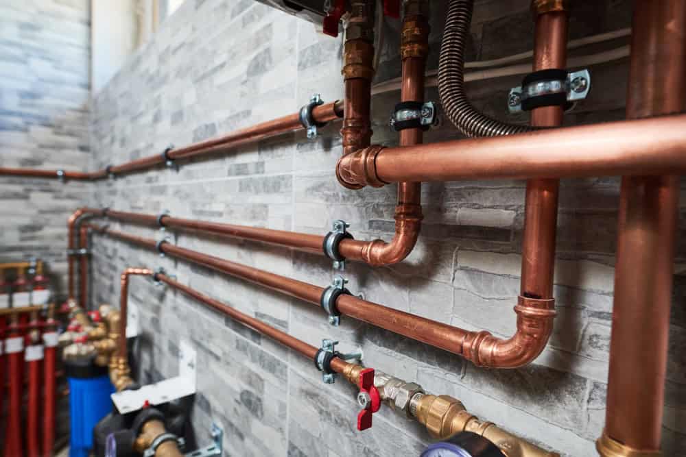 Copper pipes, expertly installed by a skilled plumber, line the gray brick wall of a mechanical room. Featuring valves and joints, this well-organized plumbing system is supported by metal clamps. Ideal for boiler services or oil to gas conversions, it reflects precision and efficiency.