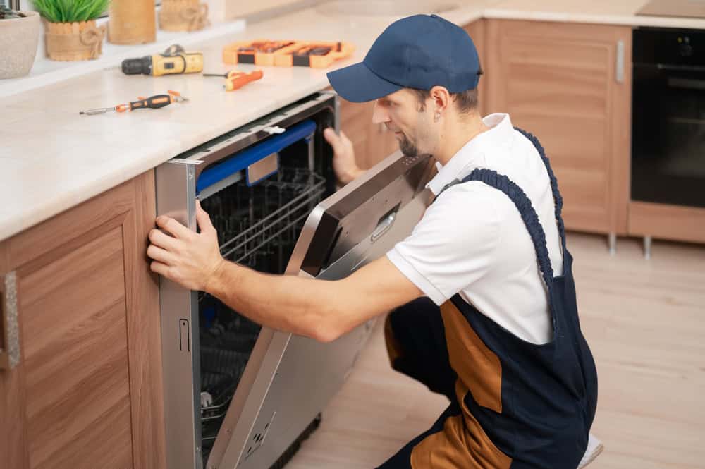 A technician wearing overalls and a cap is kneeling in front of an open dishwasher, inspecting it. Various tools are placed on the kitchen counter nearby, similar to those used in boiler services. The setting is a modern kitchen with wooden cabinets and light-colored flooring.