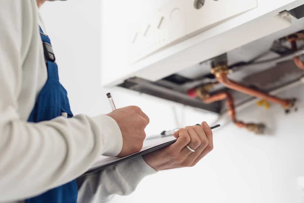 A person in a blue uniform inspects a boiler, providing expert boiler services. They hold a clipboard and pen for taking notes, focusing on the copper pipes and components of the heating system.