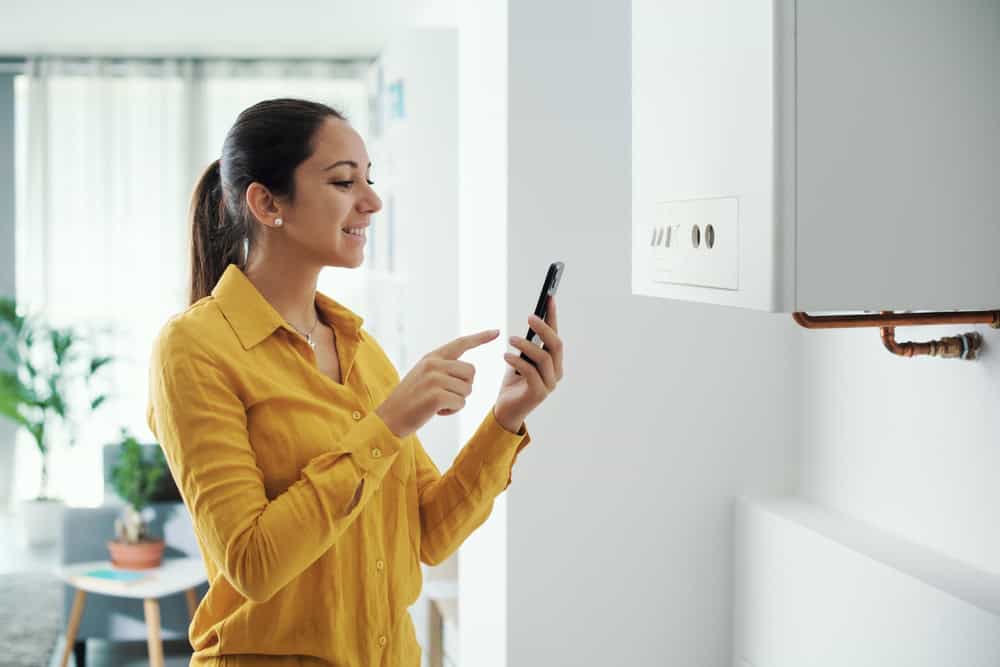 A woman in a yellow shirt stands smiling in a bright room, using a smartphone. She seems to be adjusting her Mini Split AC installation or interacting with a smart thermostat on the wall. A potted plant is blurred in the background, complementing the tech-savvy ambiance.