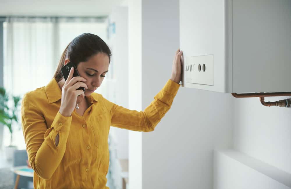 A woman in a yellow shirt is talking on her phone, likely consulting a residential plumber, while standing beside a boiler in a modern, well-lit room. She looks concerned and has her other hand resting on the boiler.