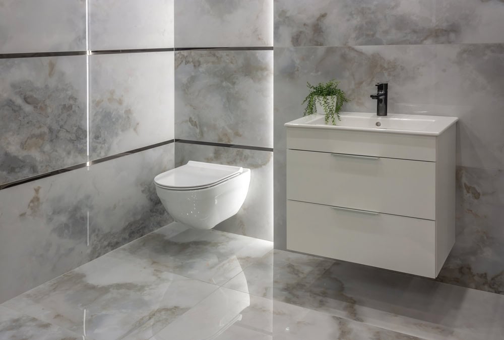 A modern bathroom featuring a wall-mounted toilet and a white vanity with two drawers. The basin has a black faucet, complemented by a small potted plant on top. With light gray marbled tiles and expert craftsmanship, the space is perfect for those considering plumber services or mini split AC installations.