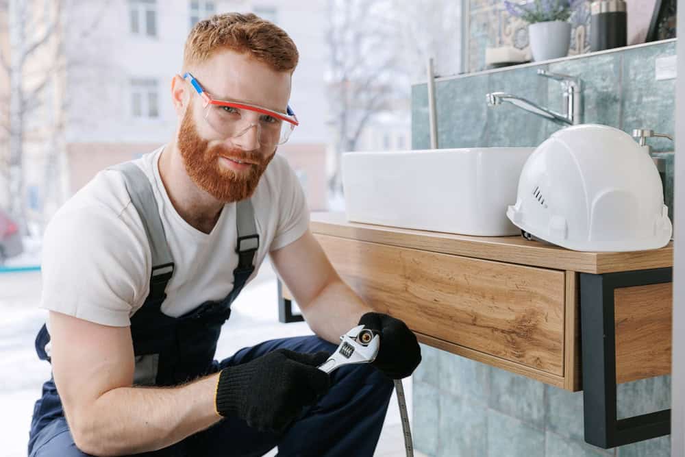 A bearded residential plumber, wearing safety glasses and gloves, is crouched down with a wrench, skillfully working on a bathroom sink. A white hard hat rests on the wooden cabinet beside him.