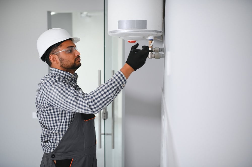 A residential plumber wearing safety gear and a white hard hat inspects a wall-mounted water heater. Dressed in a plaid shirt, overalls, and gloves, they focus intently on the task at hand against a plain interior wall.