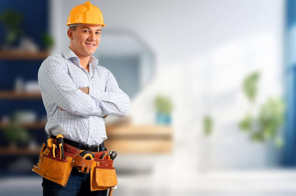 A construction worker wearing a hard hat and plaid shirt smiles confidently with arms crossed, his tool belt ready for action. The blurred indoor setting with plants and shelves subtly hints at his expertise in Mini Split AC installations.