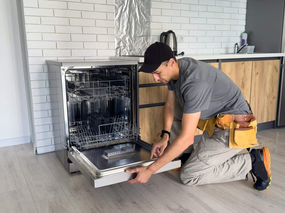 A residential plumber wearing a cap and tool belt kneels in front of an open dishwasher, inspecting or repairing it. The kitchen features white brick walls, wooden cabinets, and various appliances on the counter.