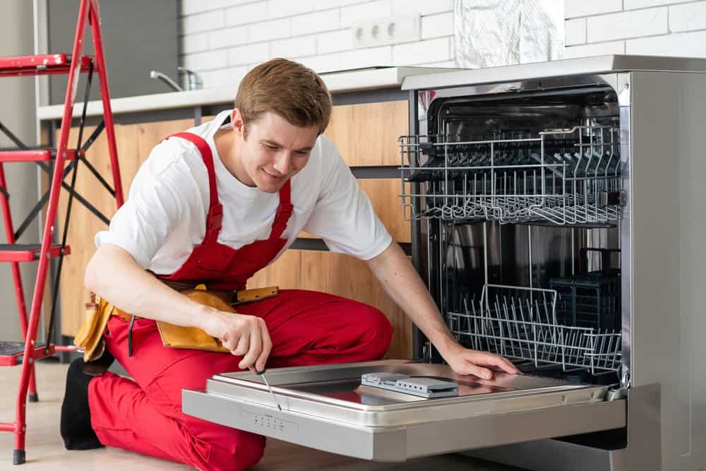 A technician in a red overall, resembling a residential plumber, is kneeling and inspecting an open dishwasher in a modern kitchen. A red stepladder stands nearby, with wooden countertops and white-tiled walls completing the scene.