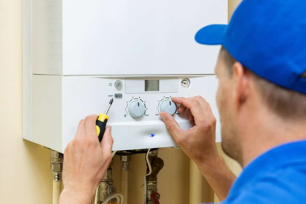 A technician in a blue uniform provides expert boiler services, adjusting the settings on a white gas boiler. They hold a screwdriver while turning the dials on the control panel, which is mounted on a beige wall, ensuring optimal performance for the household.