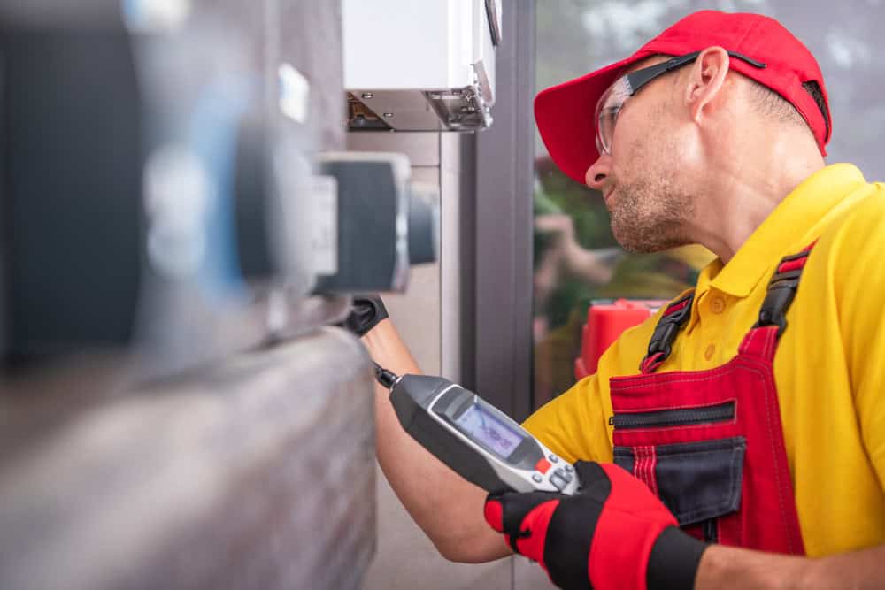 A technician in a red cap and overalls uses a handheld diagnostic device to inspect machinery. Like a meticulous residential plumber, he is focused on his work, surrounded by mechanical equipment, wearing protective eyewear and gloves.