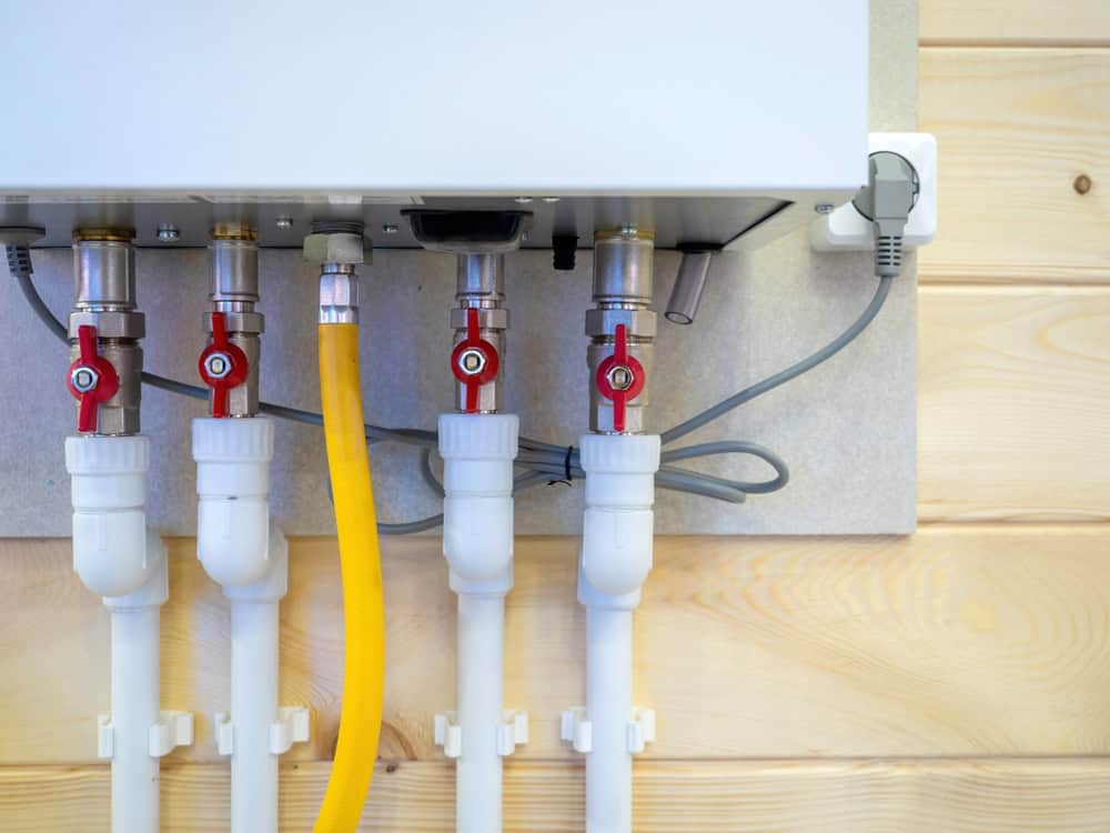 Close-up of a tankless water heater installed on a wooden wall, showcasing its efficient design. Five pipes with red valves and one yellow hose connect to the bottom, perfect for homes transitioning from oil to gas conversions. An electrical cord is plugged into a nearby outlet.